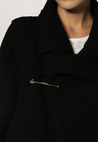 Svart strukturerad kappa med en bred krage, prydd med en framträdande silverfärgad säkerhetsnål som accent. Närbild som visar tygdetaljer och design.