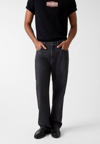 Zwarte spijkerbroek met een rechte pijp, voorzien van steekzakken aan de voorkant, een knoopsluiting en contrasterende stikseldetails. Draagt een zwart grafisch T-shirt.