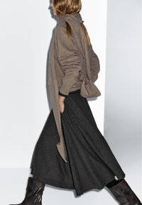 Brauner Cardigan mit drapiertem Design über einem schwarz-grau karierten Rock, kombiniert mit braunen kniehohen Stiefeln. Glatte und strukturierte Texturen.