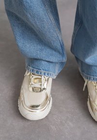 Le sneaker presentano una tomaia in rete beige con accenti in oro metallico, lacci bianchi e una suola bianca spessa e testurizzata. Indossate con jeans azzurri chiari.