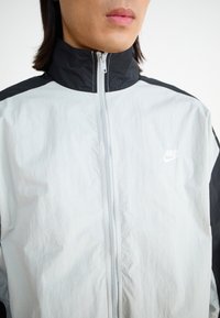 Nike jakna z visokim ovratnikom, zadrgo spredaj in svetlo sivo telo z črnimi detajli na ramenskem in rokavskem delu. Teksturno blago.