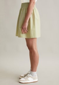 Lichtgroene, knielange rok met een smock tailleband. Heeft een zachte textuur en valt recht. Draagt met witte en beige sneakers.