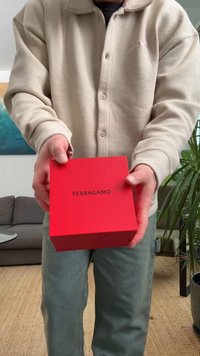Rdeča kvadratna darilna škatla z črnim napisom "FERRAGAMO" na vrhu, ki jo drži oseba, oblečena v bež teksturirano srajco in svetlo zelene hlače.