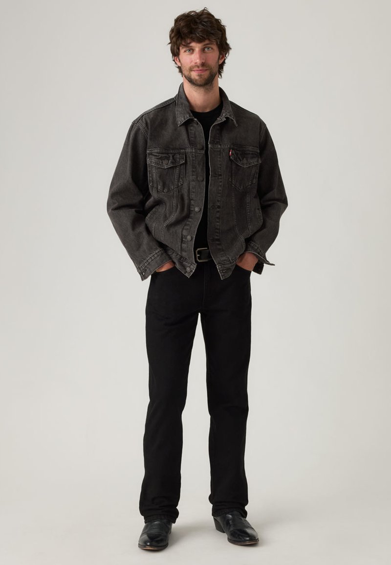 Giacca di jeans nera con due tasche sul petto, indossata sopra una camicia nera. Abbinata a pantaloni aderenti neri e scarpe nere, in piedi su uno sfondo neutro.