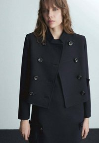 Black cropped jacket with a high collar, double-breasted button closure, and textured fabric, paired with a matching dress featuring pockets.