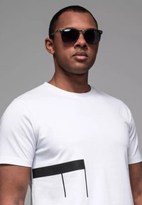 Camiseta blanca con un diseño geométrico negro en el lado izquierdo, usada con gafas de sol oscuras. Tejido suave y forma ajustada.