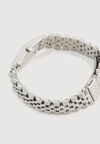 Sidabrinis metalo apyrankės laikrodis su apvaliu korpusu ir reguliuojamo ilgio grandinėle. Yra lygi poliruota apdaila ir saugus sagties uždarymas.