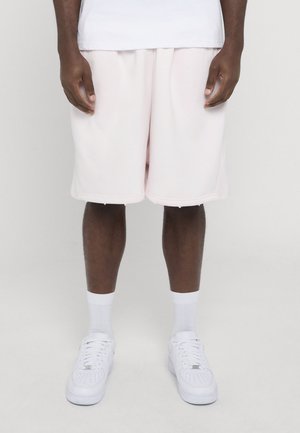 Dunkelhäutige Person trägt lockere hellrosa Shorts, weiße Crew-Socken und weiße Sneakers und steht vor einem einfarbigen hellen Hintergrund.