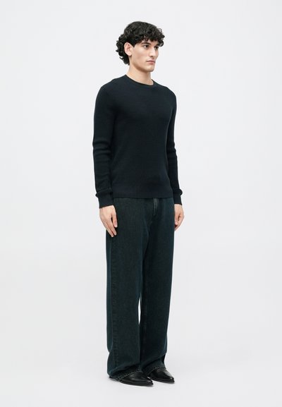 Jersey de punto negro con textura y mangas largas, combinado con jeans de denim oscuro de pierna ancha y zapatos negros de puntera, exhibido en un modelo.