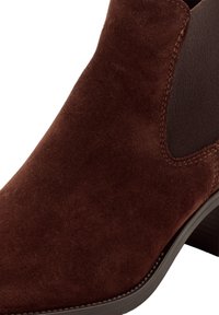 Stivaletto alla caviglia in pelle scamosciata color castagna con texture liscia, pannelli laterali elastici e tacco basso a blocco. Dettagli cuciti lungo i bordi.
