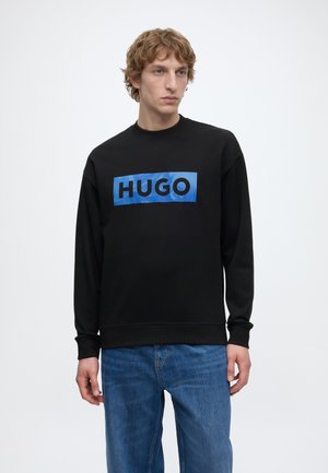 Νέος άνδρας φοράει μαύρο φούτερ με μπλε ορθογώνιο λογότυπο που γράφει "HUGO" και μπλε τζιν, στέκεται μπροστά σε απλό φόντο.