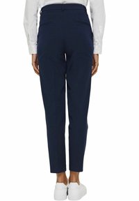 Vue de dos d'une personne portant un pantalon slim bleu marine, une chemise blanche à manches longues et des baskets blanches, debout sur un fond blanc.