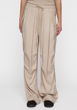 Pantaloni beige a vestibilità ampia con elastico in vita e tasche laterali indossati con scarpe scure su una persona su uno sfondo neutro.