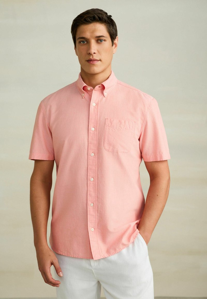 Homme portant une chemise à manches courtes rose clair avec une poche poitrine et un pantalon blanc, debout avec une main dans la poche.