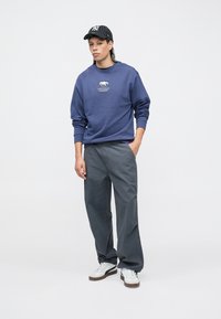 Blue sweatshirt with bear graphic, gray loose-fit pants, and white sneakers. The model wears a black and white striped cap.