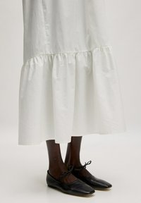 Wit, knie-lange katoenen jurk met een gegatherde zoom, gecombineerd met zwarte platte schoenen met een strikdetail. Soepele stoftextuur, eenvoudig ontwerp.