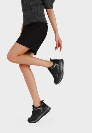 Donna che indossa sneakers platform nere, pantaloni corti neri e un top a maniche corte grigio scuro, alzando una gamba contro uno sfondo chiaro e semplice.