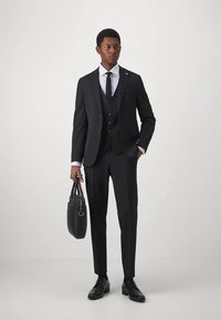 Άνδρας με μαύρο τρι-piece κοστούμι και γραβάτα κρατώντας μαύρη δερμάτινη βαλίτσα, στέκεται μπροστά σε λευκό φόντο.