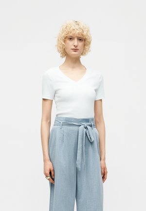 Νέα γυναίκα με σγουρά ξανθά μαλλιά που φοράει λευκό κοντομάνικο μπλουζάκι με V λαιμόκοψη και ανοιχτό μπλε ψηλόμεσο παντελόνι με δεμένη ζώνη.