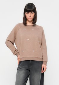Sweatshirt marron avec des accents de motif argent, col rond et manches raglan. Porté avec un jean gris foncé, présentant une coupe décontractée et une texture.