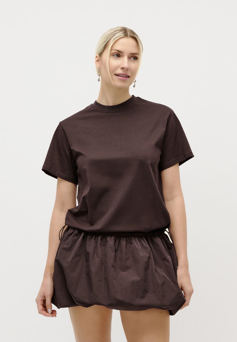 Γυναίκα που φοράει κοντομάνικο σκούρο καφέ πουκάμισο και αντίστοιχη μίνι φούστα με πιέτες, κοιτάζει προς τα δεξιά της μπροστά σε απλό ανοιχτό φόντο.