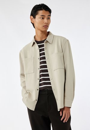 Camisa beige clara de botón con dos bolsillos en el pecho, llevada sobre una camiseta de manga larga a rayas oscuras. Presenta un corte relajado y una textura suave.