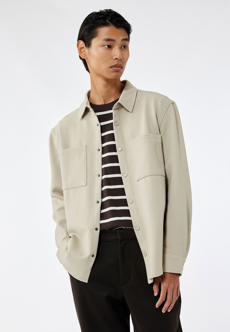 Camisa beige clara de botón con dos bolsillos en el pecho, llevada sobre una camiseta de manga larga a rayas oscuras. Presenta un corte relajado y una textura suave.