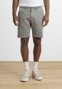 Shorts verts texturés avec fermeture à bouton et poche frontale. Manches roulées. Associés à des baskets beiges et des chaussettes blanches de type crew.
