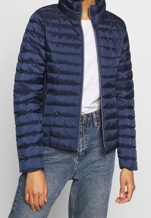 Person iført en navyblå quiltet jakke over en hvid skjorte og blå jeans, stående mod en ensfarvet baggrund.