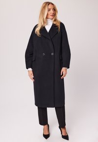 Długi czarny wełniany płaszcz z klapowym kołnierzem, podwójnym zapięciem na guziki z przodu i bocznymi kieszeniami, noszony na czarnych spodniach i szpilkach.