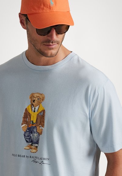 Gaiši zils T-kreklis ar attēlu, kurā attēlots lācītis brūnā jakā, dzeltenā šallē un džinsos, ar tekstu "POLO BEAR by RALPH LAUREN."