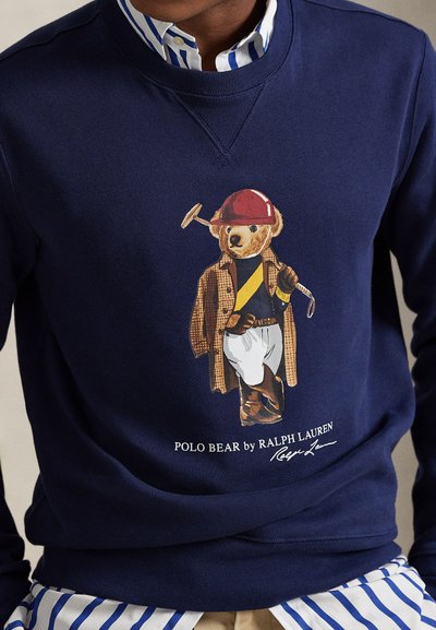 Marineblå genser med en Polo Bear som har på seg en brun frakk, rød hjelm, hvite bukser, holder en polomalk, lag på lag med en stripete skjorte.