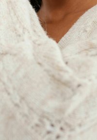 Pull en tricot couleur crème avec un motif texturé, offrant une apparence douce et confortable, et un design en V. Gros plan sur les détails du tissu.