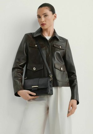 Kobieta w ciemnobrązowej skórzanej kurtce i białych spodniach, trzymająca czarną torbę na pasku z złotym zapięciem na jasnym tle.