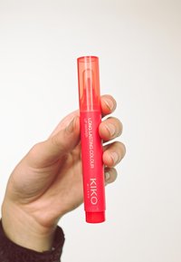Red lip marker with transparent cap, featuring bold white lettering, round shape, and smooth texture, held between fingers against a light background.