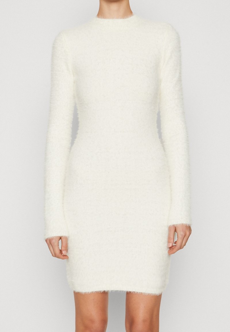 Femme portant une robe pull blanche moulante à manches longues et longueur genou, en tissu duveteux, sur un fond clair uni.