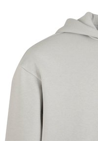 Sweat à capuche gris clair à manches longues, coupe ample, capuche visible et texture de tissu lisse, vu depuis la zone supérieure de l'épaule.