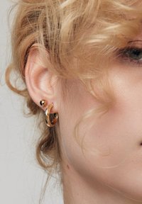 Złote kolczyki nausznice o młotkowanej fakturze, uzupełnione małym złotym wkrętem. Włosy są delikatnie pokręcone, otaczając ucho.