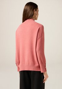 Maglione rosa lavorato a maglia con collo alto, vestibilità ampia e decorazioni a bottoni sulle maniche. Texture morbida e design informale.