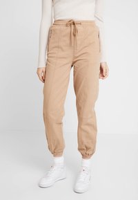 Beżowe spodnie dresowe wykonane z lekkiego materiału, z elastycznym pasem ze sznurkiem, bocznymi kieszeniami na zamek błyskawiczny oraz zwężanymi mankietami.