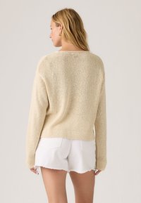 Γυναίκα με ξανθά μαλλιά, ντυμένη με μπεζ πλεκτό πουλόβερ και λευκά τζιν σορτς με σκισίματα, που στέκεται μπροστά σε απλό ανοιχτό φόντο.