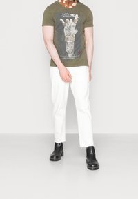 T-shirt graphique vert olive avec un design de main, associé à un pantalon court blanc et des bottes de cheville noires avec des accents métalliques.