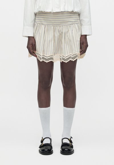 Personne portant une jupe courte rayée avec bordure en dentelle, une chemise blanche à manches longues, des chaussettes blanches et des chaussures noires à boucle, debout devant un fond uni.