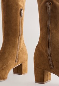 Hellbraune kniehohe Wildlederstiefel mit Blockabsatz, ausgestattet mit zwei seitlichen Reißverschlüssen und einer weichen Textur. Das Design beinhaltet Zierstiche.