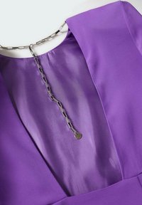 Robe violette avec un col en satin et un collier en chaîne argentée à maillons rectangulaires et un pendentif rond sur un fond clair.