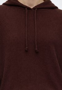 Gros plan d'un sweat à capuche marron texturé avec des cordons noués aux extrémités, porté par une personne.