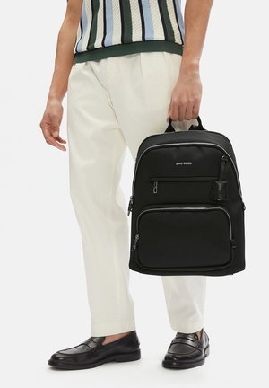 Uomo che indossa pantaloni color crema e mocassini neri che tiene uno zaino nero Gino Rossi con tasche frontali con cerniera.