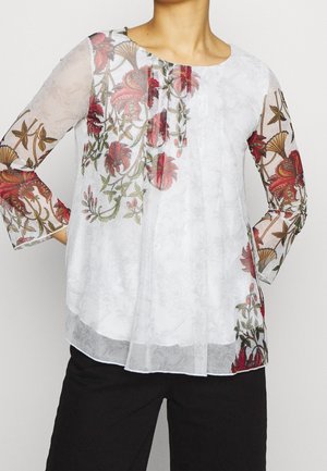 Blouse blanche en tissu transparent avec broderies florales rouges et vertes sur les manches et le haut de la poitrine, associée à un pantalon noir.