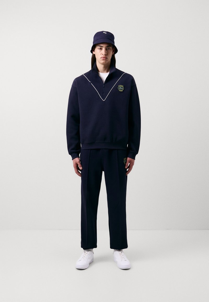 Sweatshirt marine avec zip moitié et coutures blanches, associé à un pantalon assorti. Un chapeau seau complète la tenue, avec un logo.