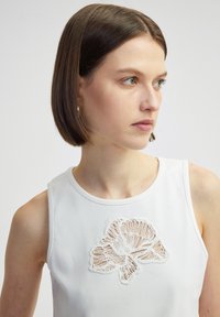 Witte mouwloze top met een gestructureerde stof en een geborduurd bloemenontwerp in het midden, met complexe naden en details.